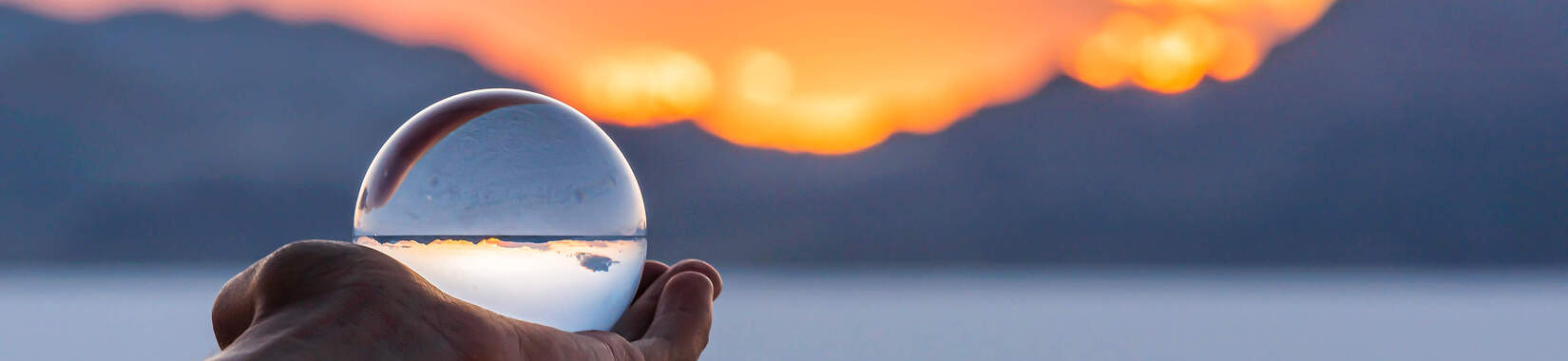 A hand holds a glass ball in front of a sunset